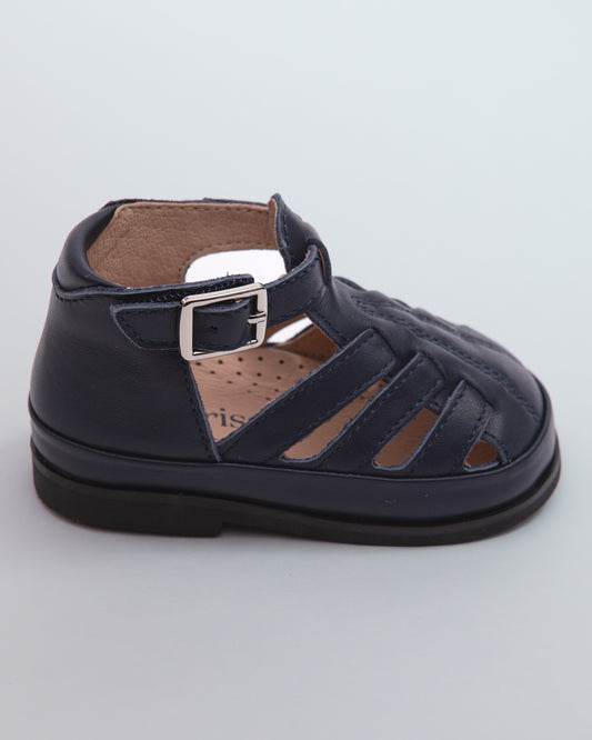 Side view of a single navy leather baby boy sandal with rubber sole, open cutouts, and velcro strap with a silver buckle, shown against a pale grey surface.