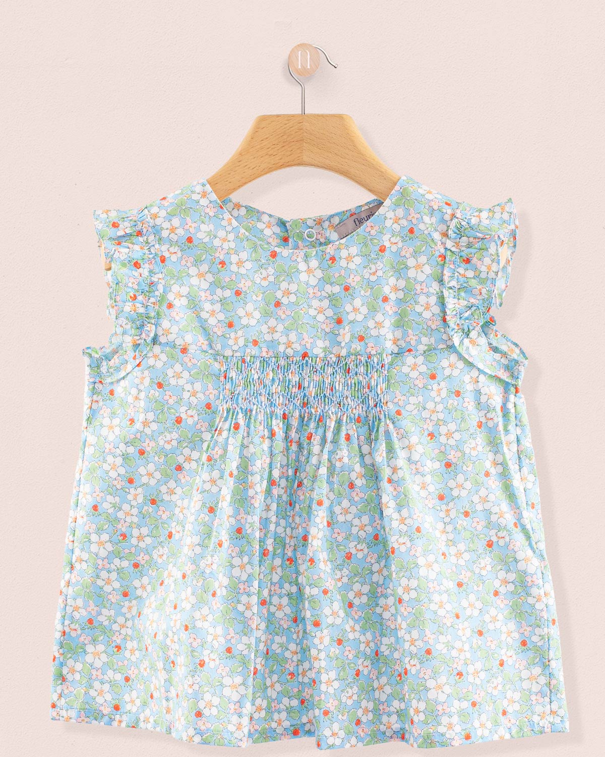 Front view of the Amelie Liberty Strawberry Field Blue Tunic, a sleeveless A-line top with hand-smocked detailing and ruffled sleeves in a pale blue floral Liberty print with white blossoms and small red strawberries.