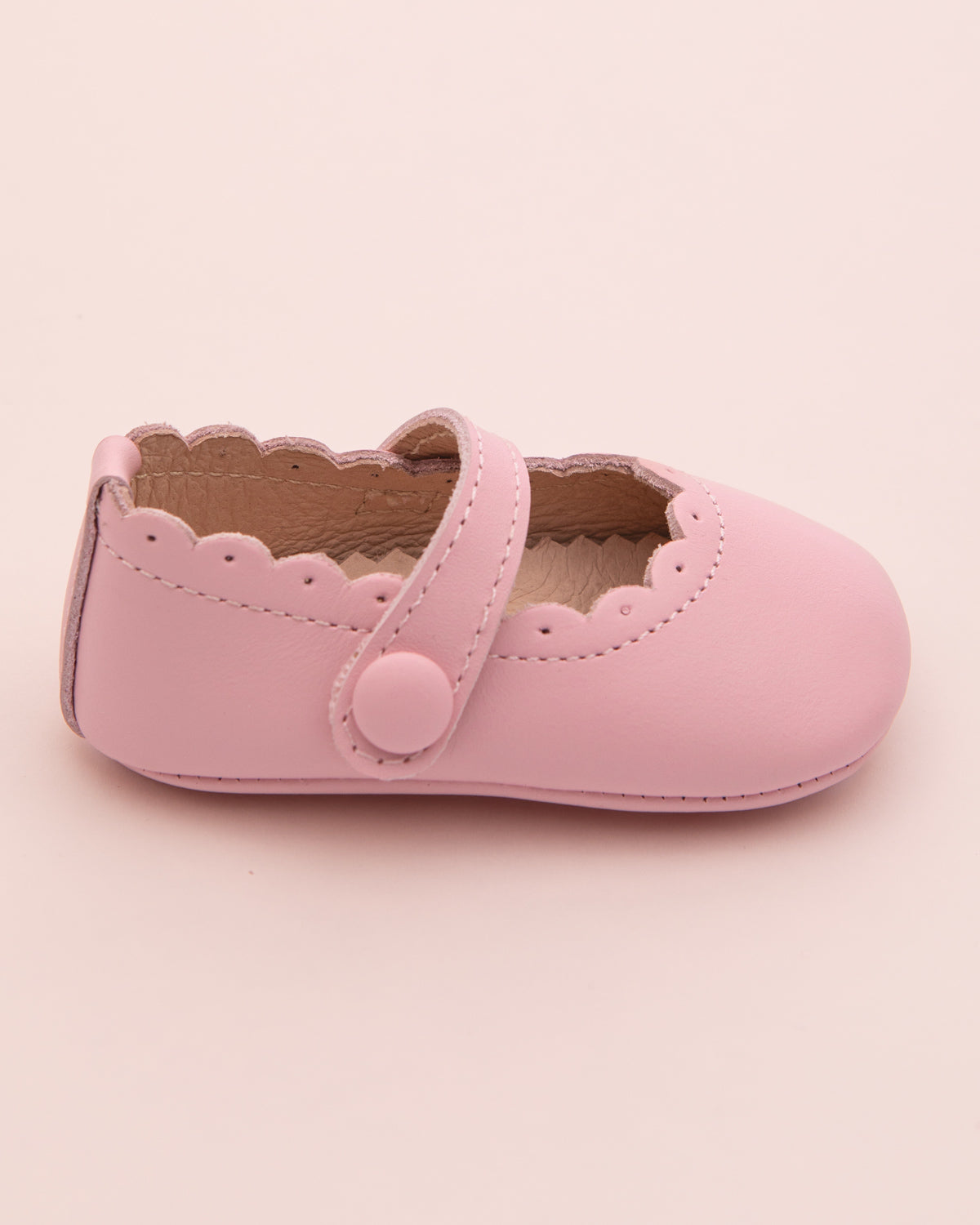 Side view of a single blush pink leather baby crib shoe with scalloped trim and velcro strap closure, shown on a pale pink surface.