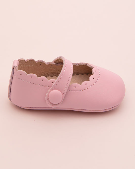 Side view of a single blush pink leather baby crib shoe with scalloped trim and velcro strap closure, shown on a pale pink surface.