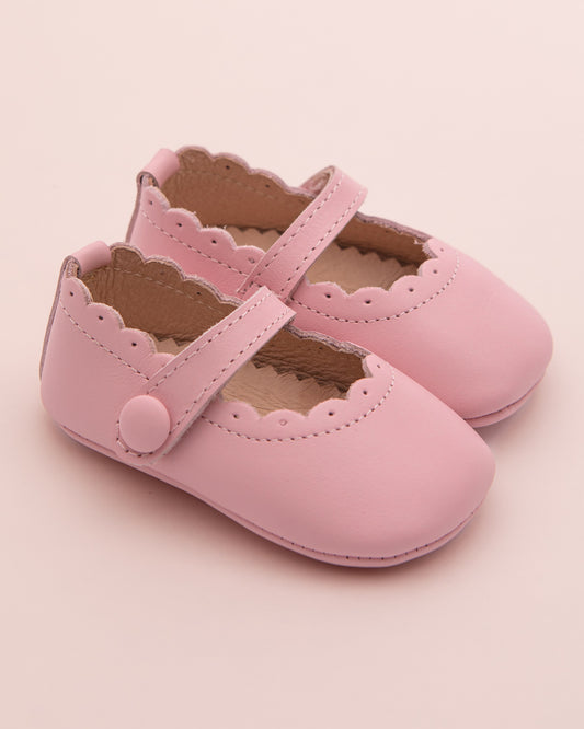 Pair of blush pink leather baby crib shoes with scalloped edges, perforated detailing, and velcro strap closure, displayed on a pale pink background.