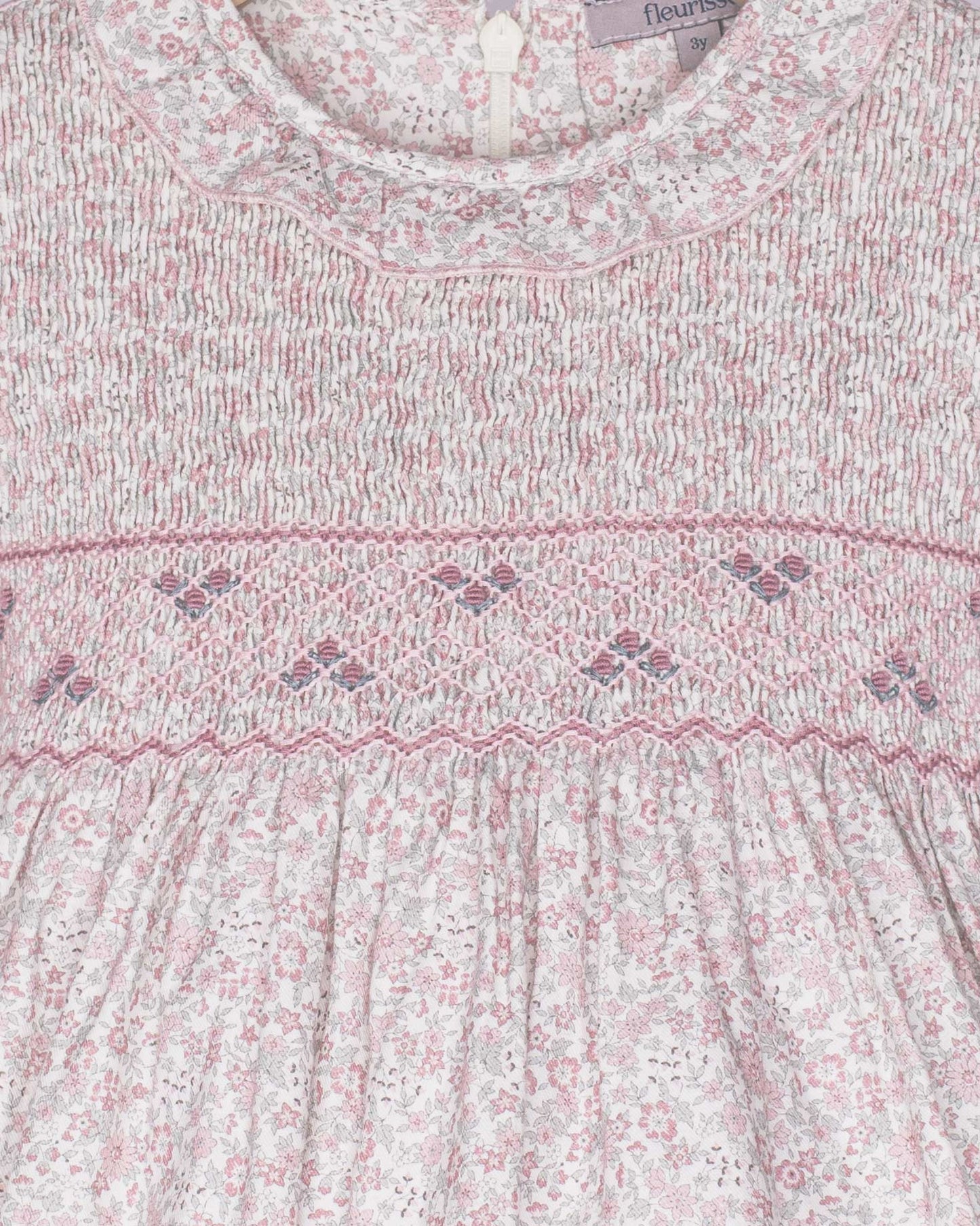 Close-up of a baby dress with floral pattern and smocked details, brand 'fleur' visible.