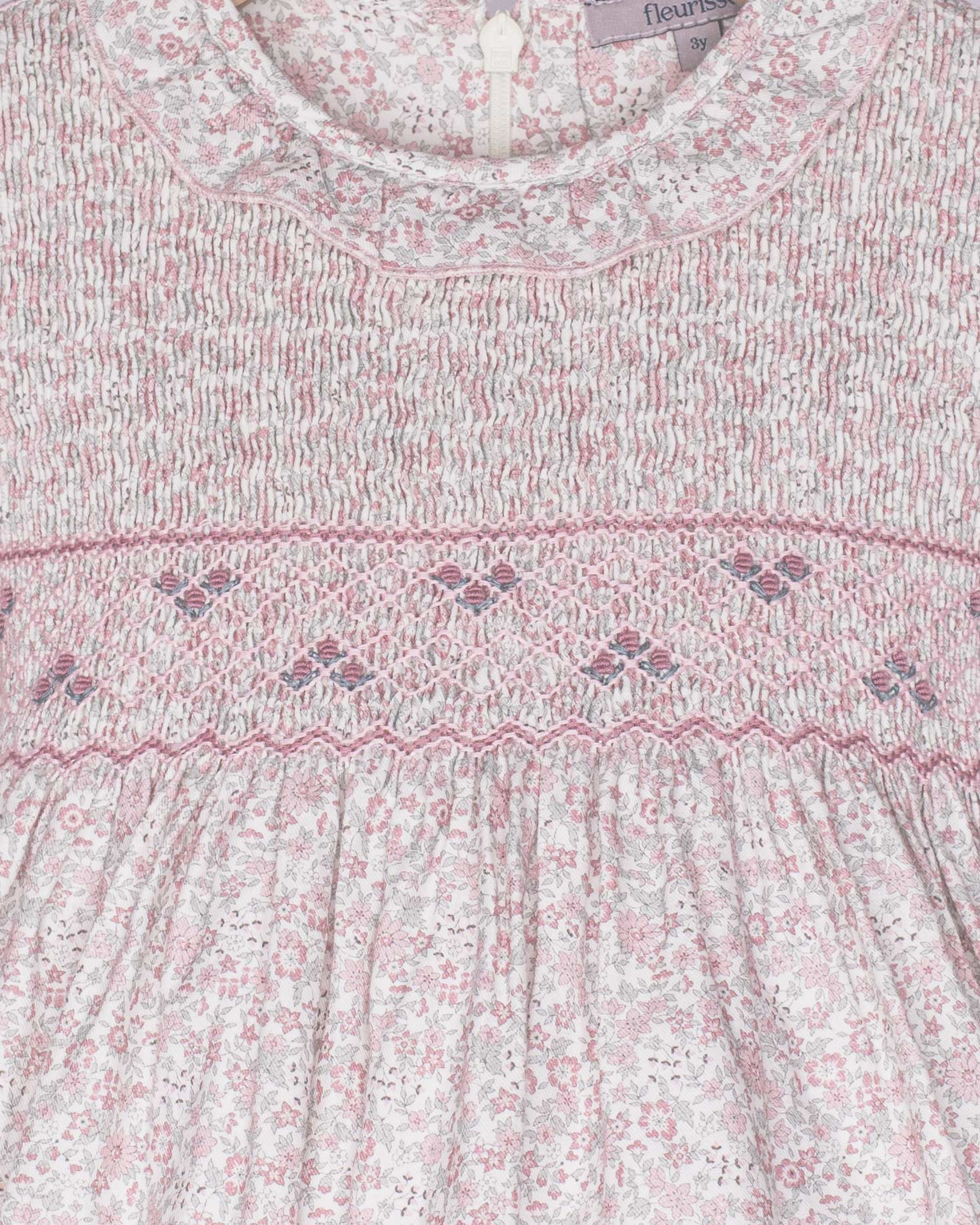 Close-up of a baby dress with floral pattern and smocked details, brand 'fleur' visible.