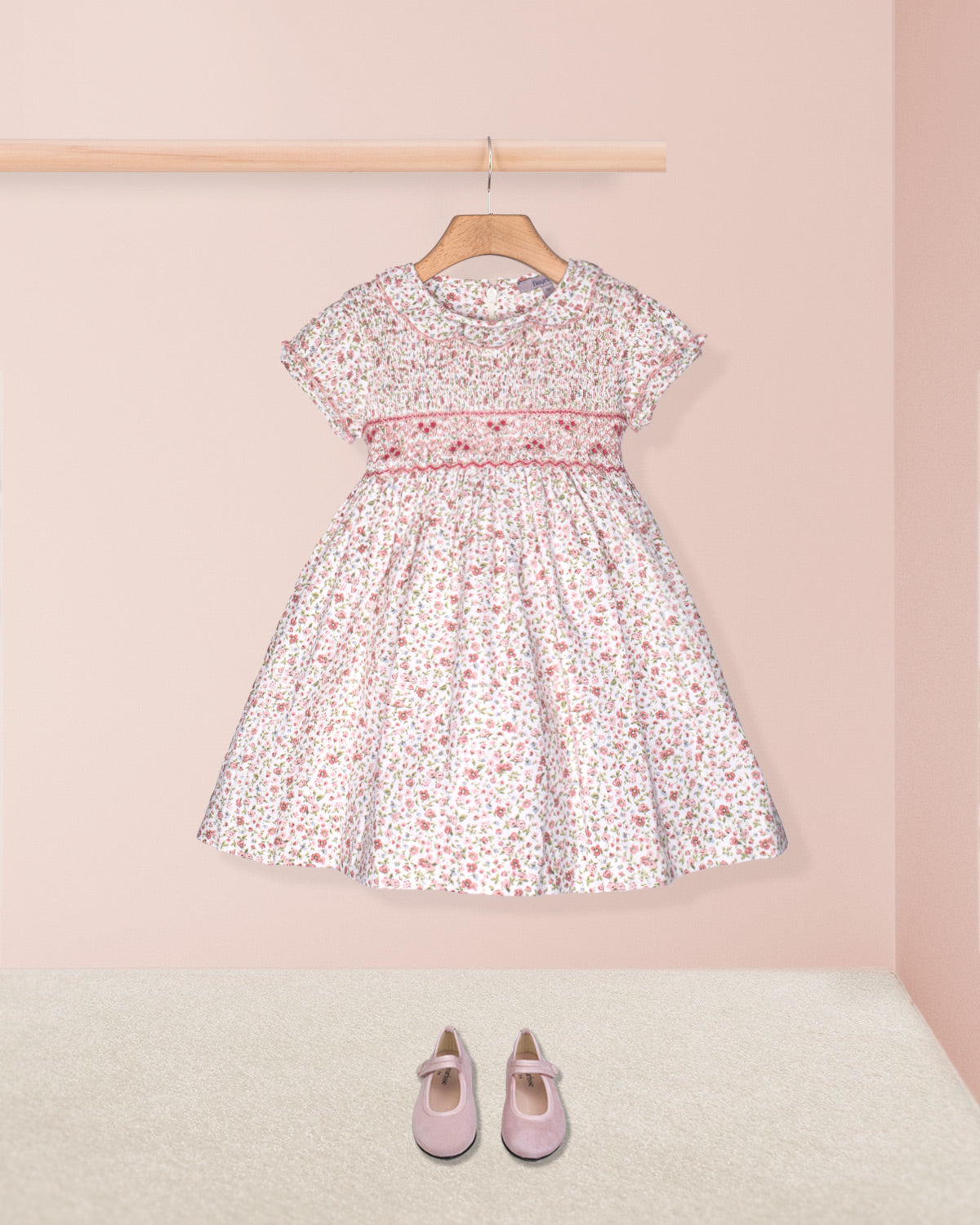 Floral dress on a hanger with pink shoes on the floor against a pink wall.