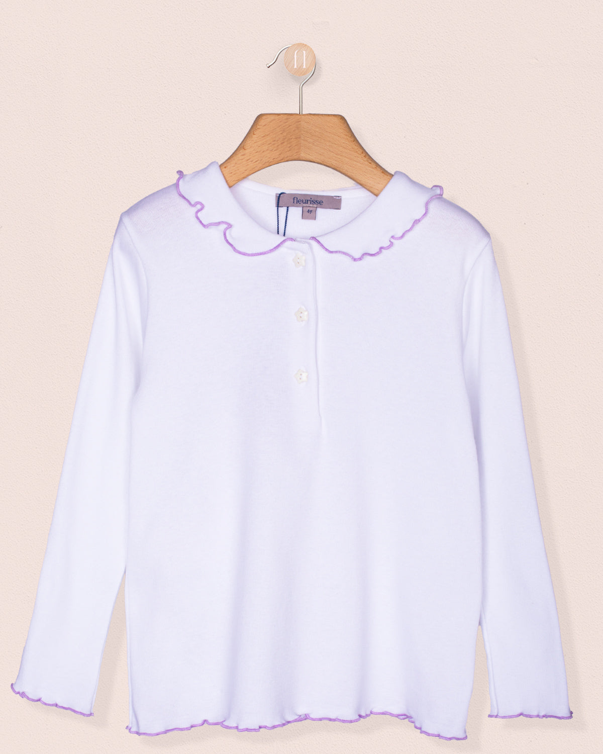 Long sleeve white brushed cotton shirt for girls displayed on a wooden hanger against a pale pink background. Features a rounded ruffle collar and hemline with lavender scalloped trim and star-shaped buttons down the front.