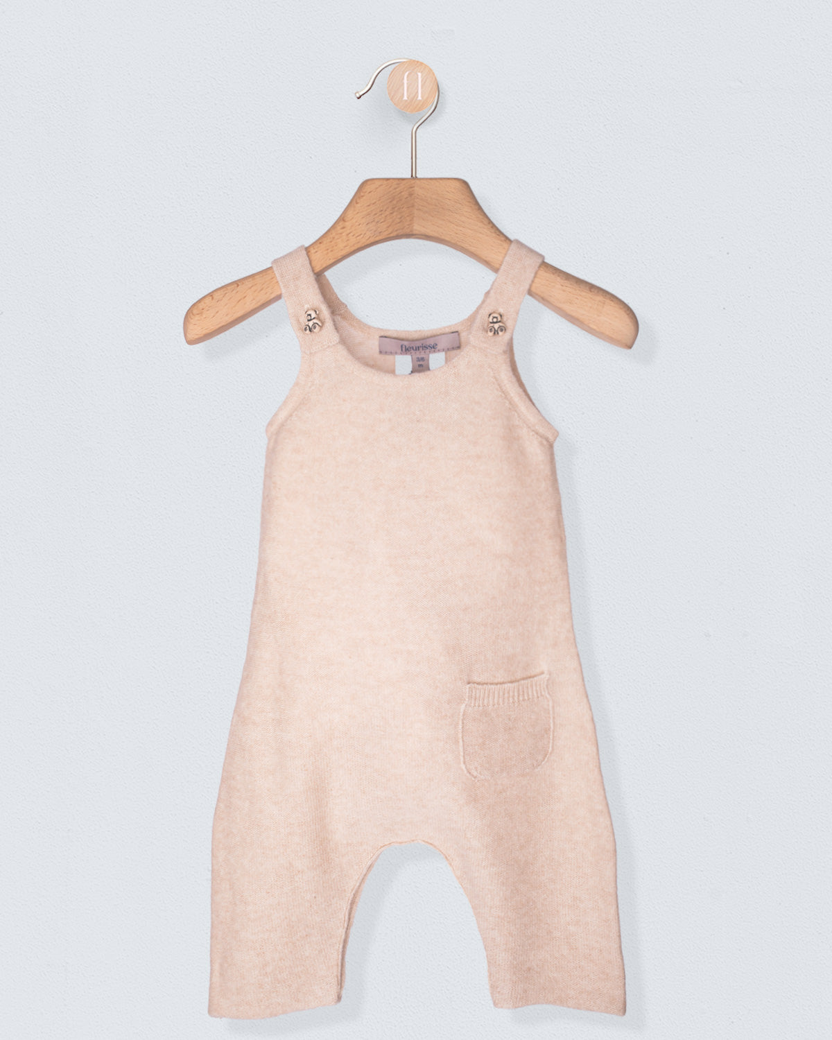 Beige cashmere overalls on a wooden hanger against a light gray background