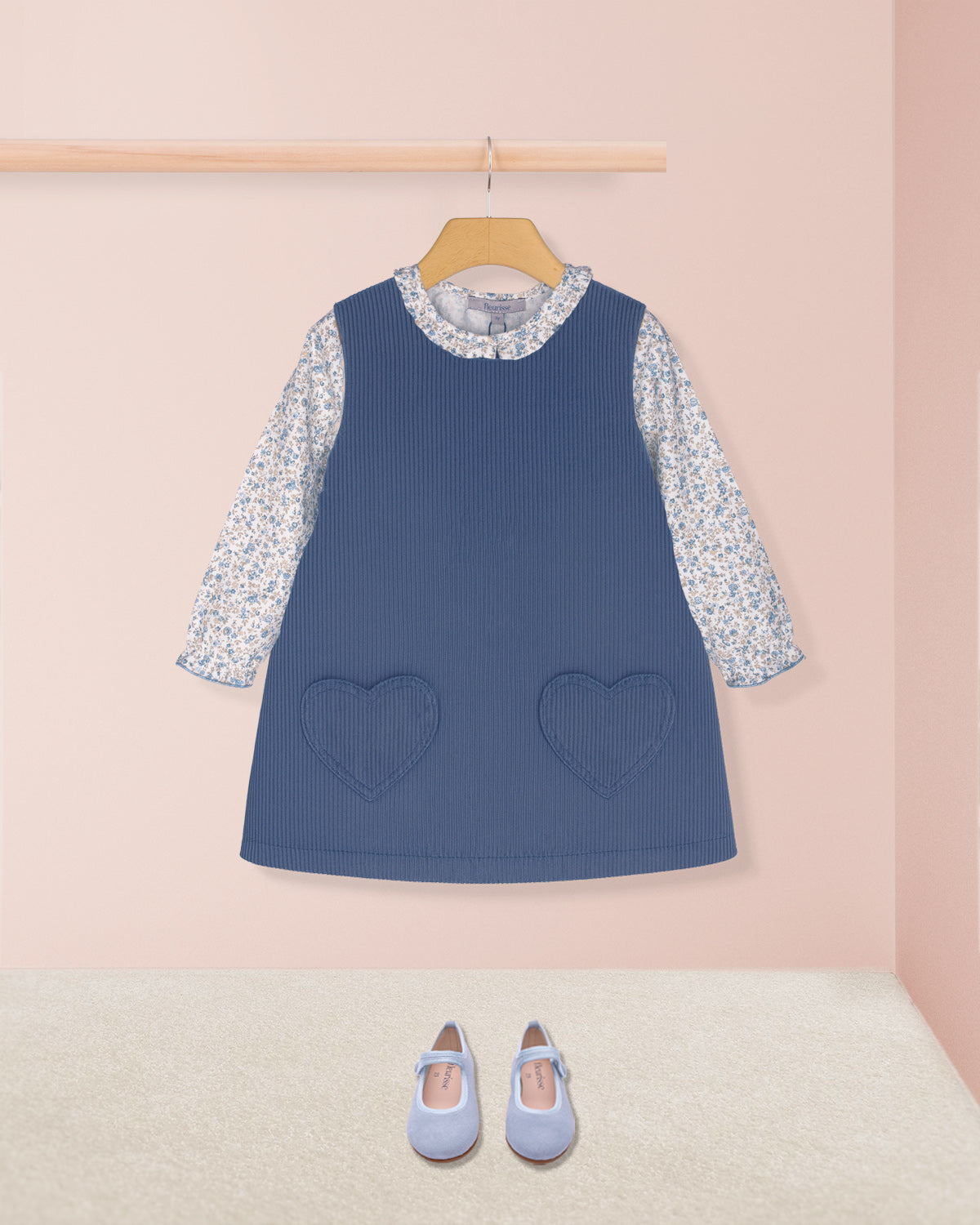 Blue dress with floral sleeves and heart pockets on a hanger against a pink wall.