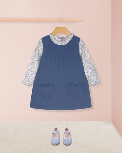 Blue dress with floral sleeves and heart pockets on a hanger against a pink wall.