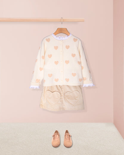 Children's outfit with heart-patterned top and skirt on a hanger against a pink wall.
