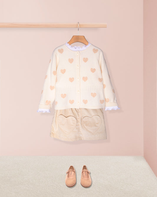 Children's outfit with heart-patterned top and skirt on a hanger against a pink wall.