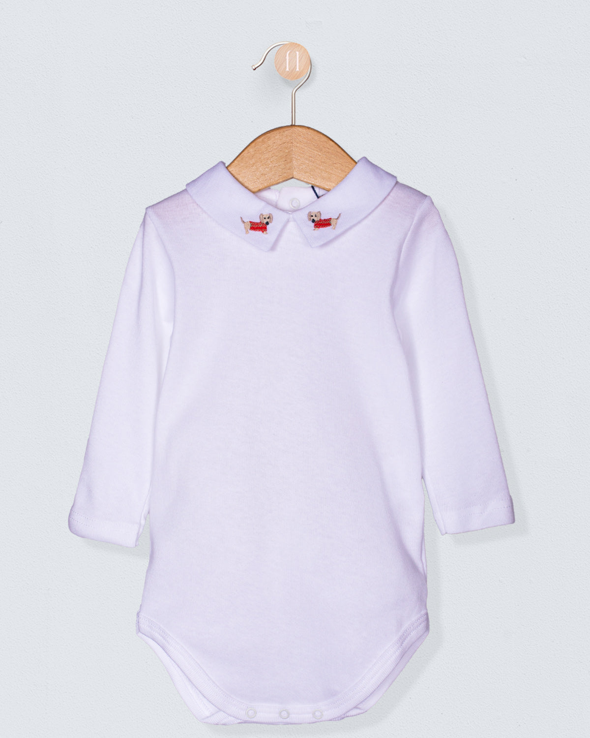 Italian cotton baby onesie with long sleeves and embroidered dachshund collar, displayed on a wooden hanger against a light background.