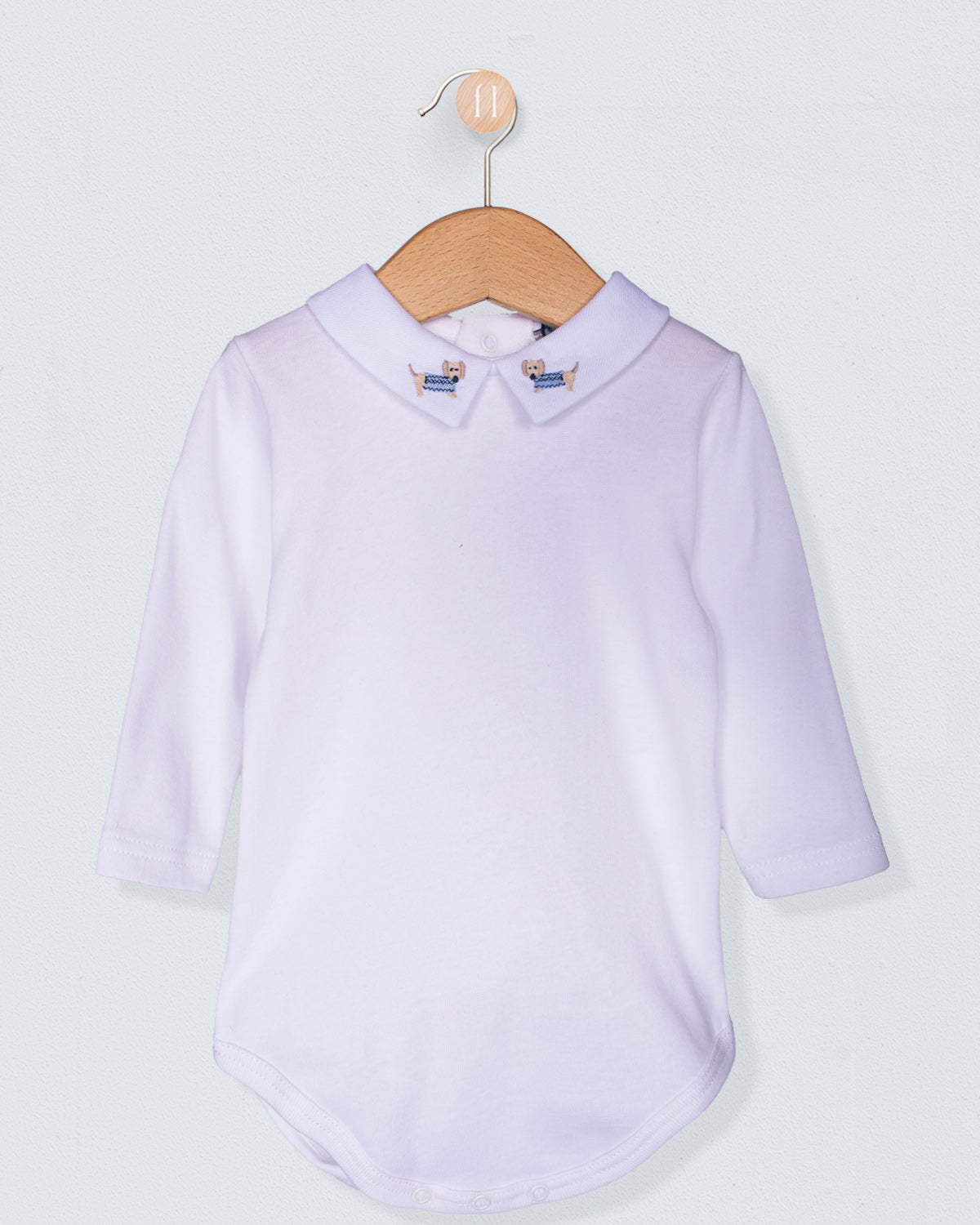 Italian cotton baby onesie with long sleeves and embroidered dachshund collar, displayed on a wooden hanger against a light background.