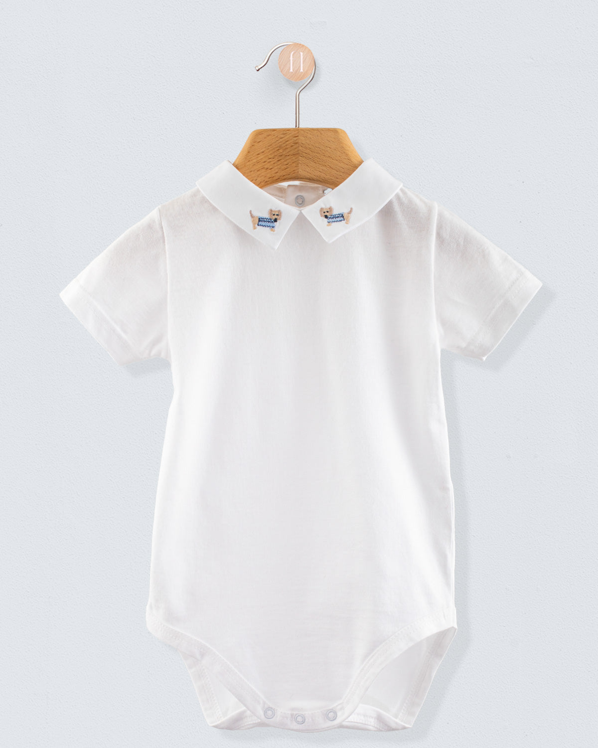 Front view of the Andreas Onesie with Embroidered Light blue Dachshund, a short-sleeve white cotton jersey onesie with a crisp collar featuring embroidered dachshund dogs in light blue sweaters.