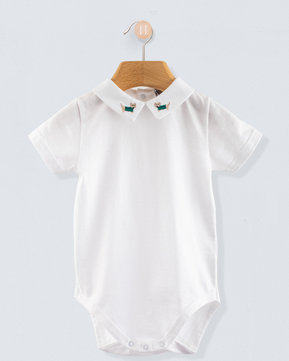 Front view of the Andreas Onesie with Embroidered Light blue Dachshund, a short-sleeve white cotton jersey onesie with a crisp collar featuring embroidered dachshund dogs in sage green sweaters.
