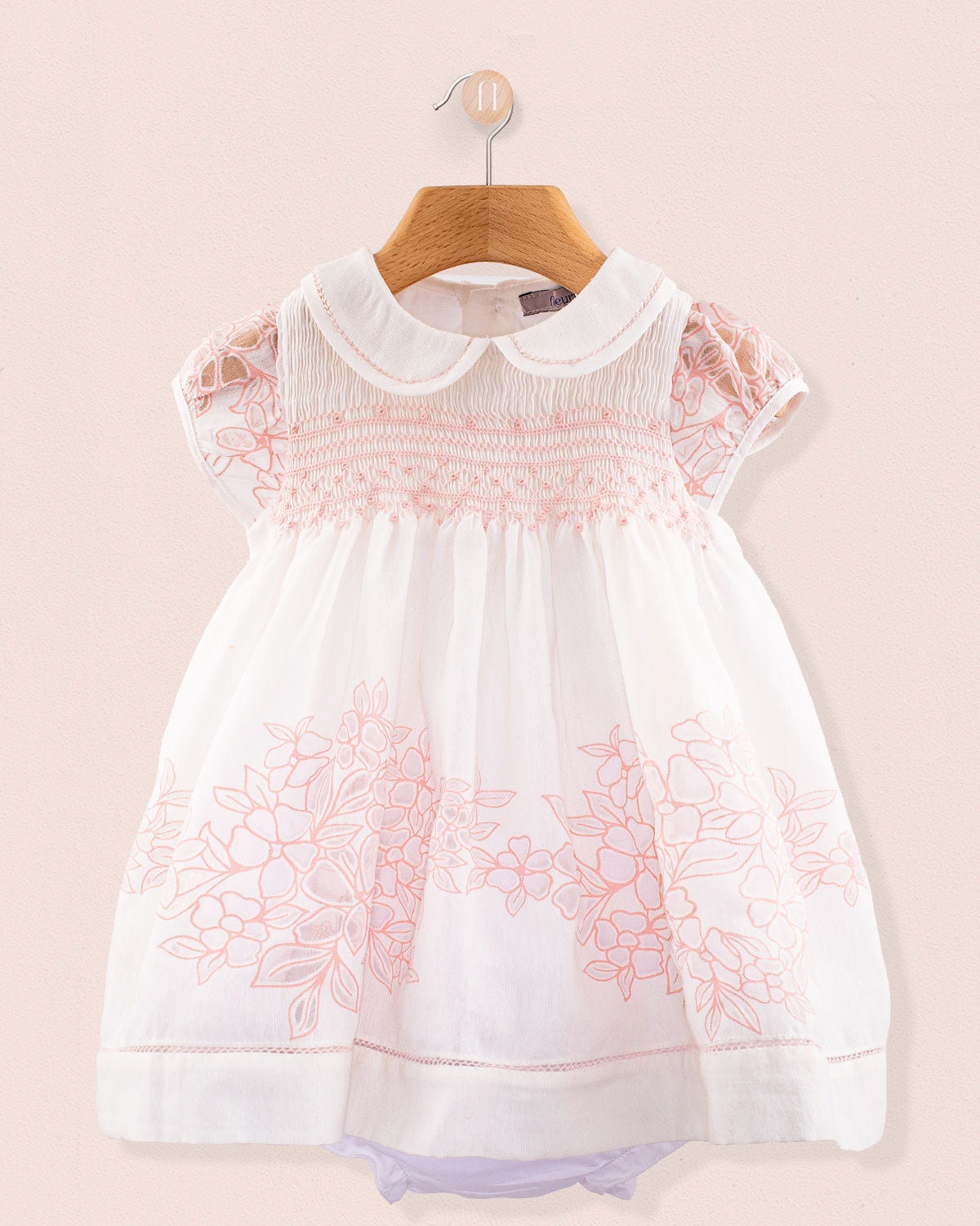 Front view of Baby Emmy Doppia Pink Smock Dress Set hanging on a wooden hanger showing pink hand smocking and floral embroidery on white cotton fabric