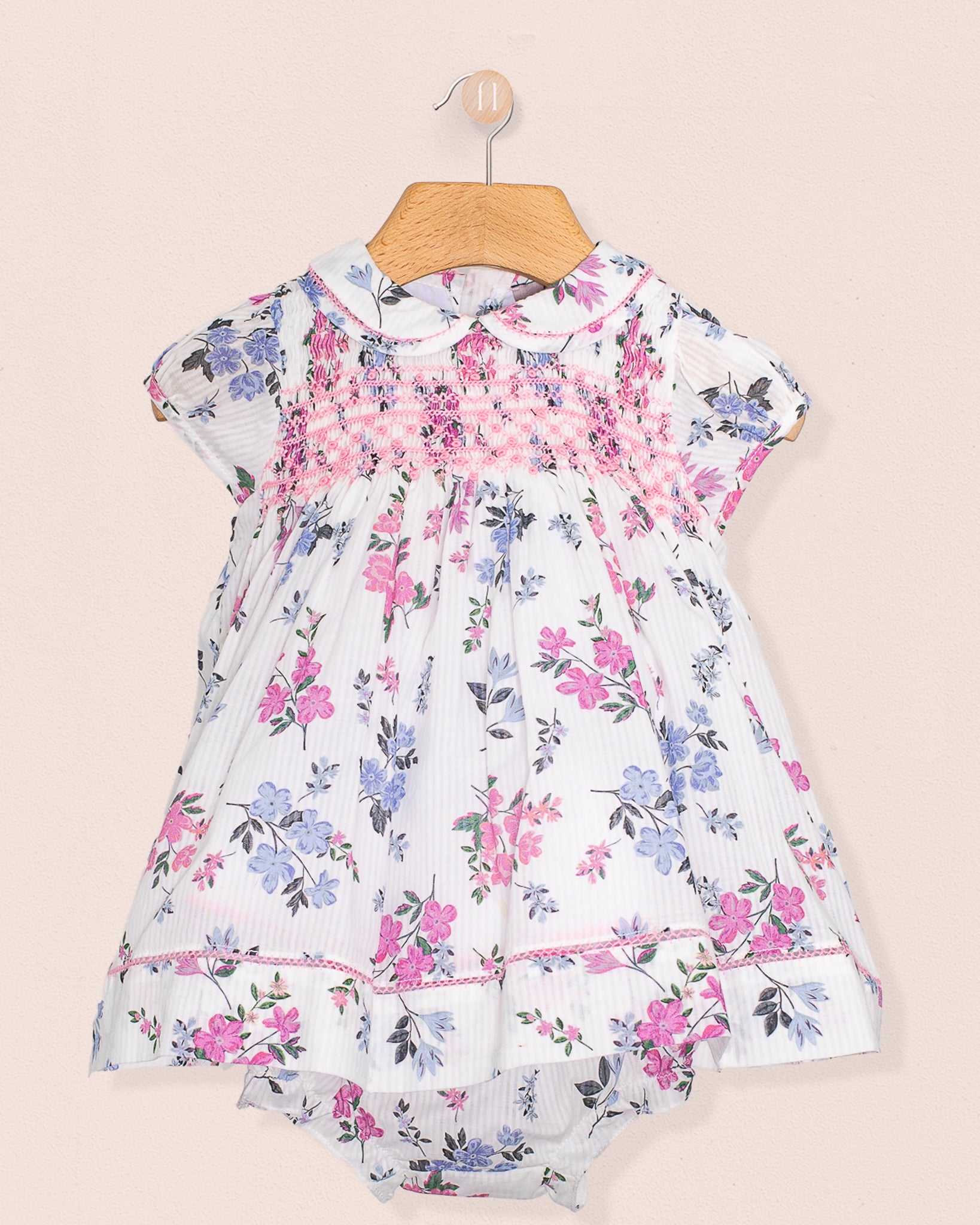A short-sleeved white baby dress with pink and blue floral print, pink smocked bodice, lace-trimmed collar and hem, paired with matching bloomers, displayed on a wooden hanger against a neutral background.