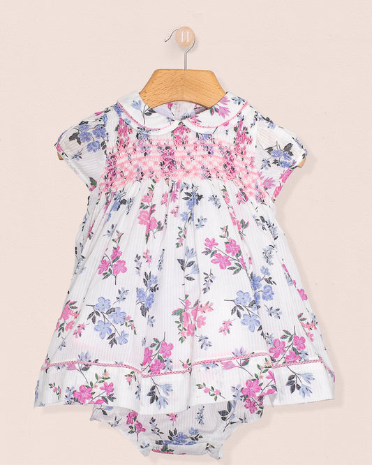 A short-sleeved white baby dress with pink and blue floral print, pink smocked bodice, lace-trimmed collar and hem, paired with matching bloomers, displayed on a wooden hanger against a neutral background.
