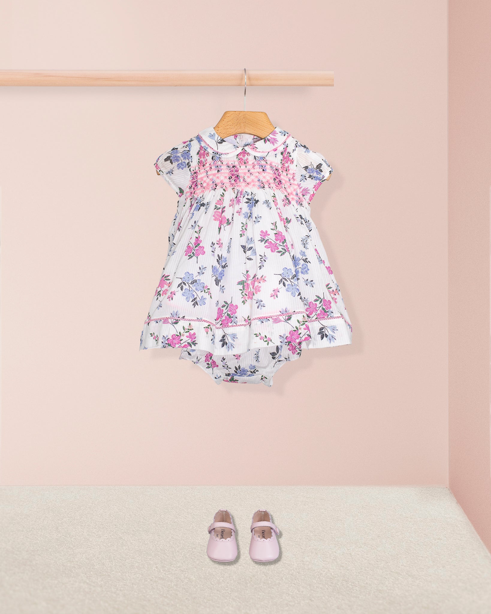 Complete outfit of Baby Emmy Whisper Dress and bloomers displayed with light pink baby shoes on carpeted floor, dress hung from wooden rail.