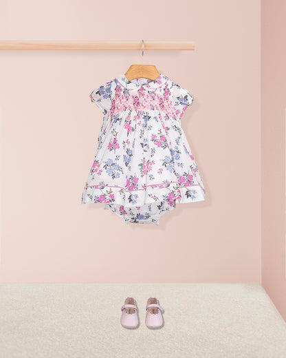 Complete outfit of Baby Emmy Whisper Dress and bloomers displayed with light pink baby shoes on carpeted floor, dress hung from wooden rail.