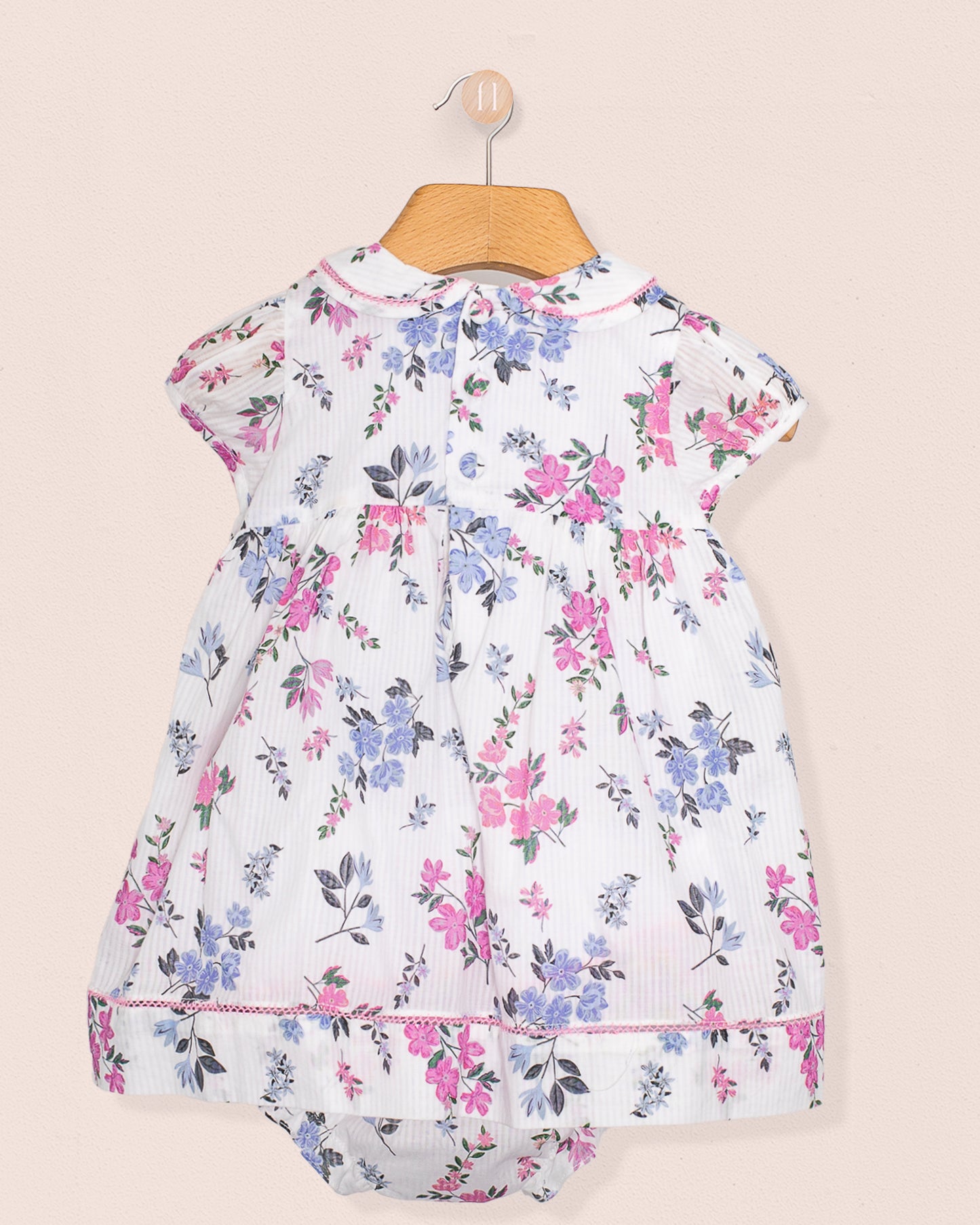 Rear view of the baby dress and bloomers showing gathered back detail and continuous floral print, hung neatly on a hanger against soft beige wall.