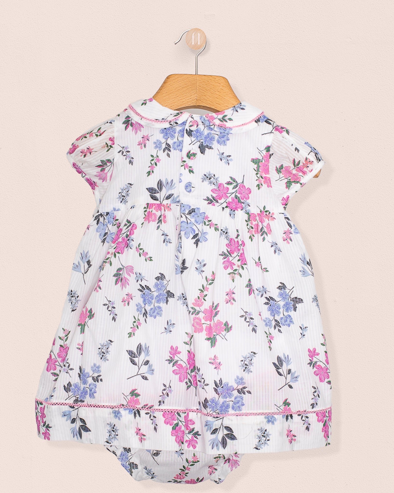 Rear view of the baby dress and bloomers showing gathered back detail and continuous floral print, hung neatly on a hanger against soft beige wall.