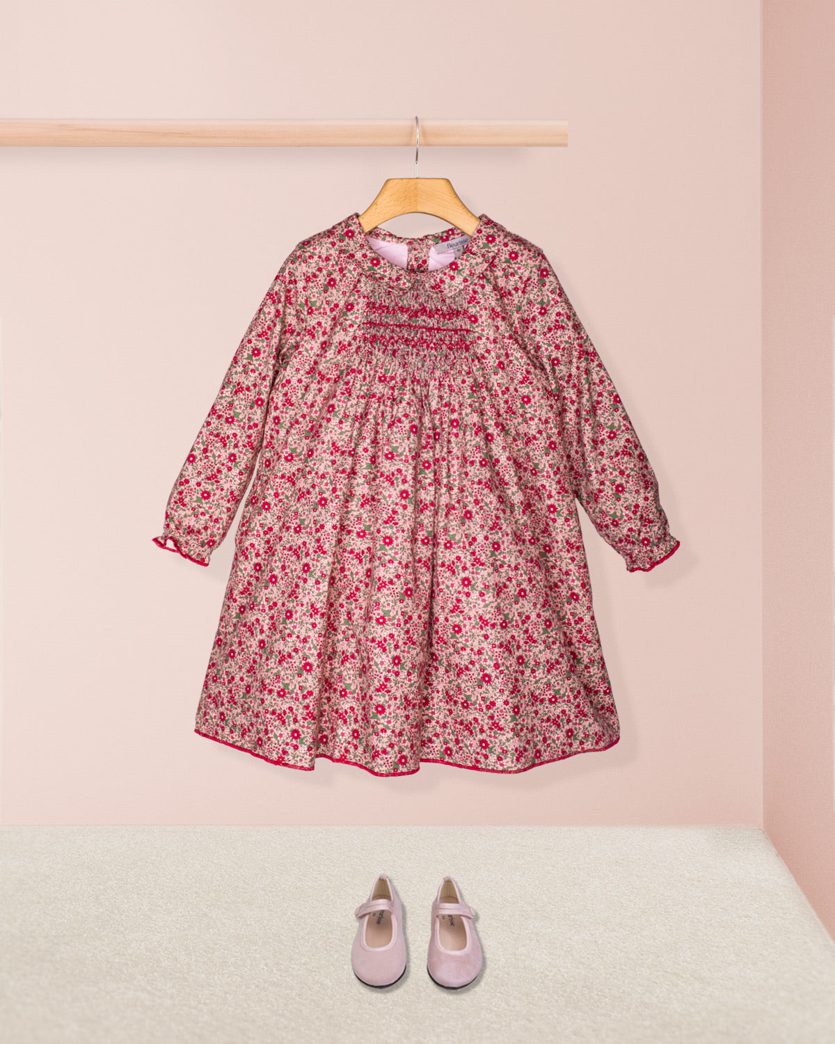 Front view of the Elizabeth Mini Mauve Floral Twill Dress hanging on a wooden hanger, showcasing its hand-smocked bodice, soft collar, and delicate mauve floral print accessorized with pink suede Mary Jane shoes.