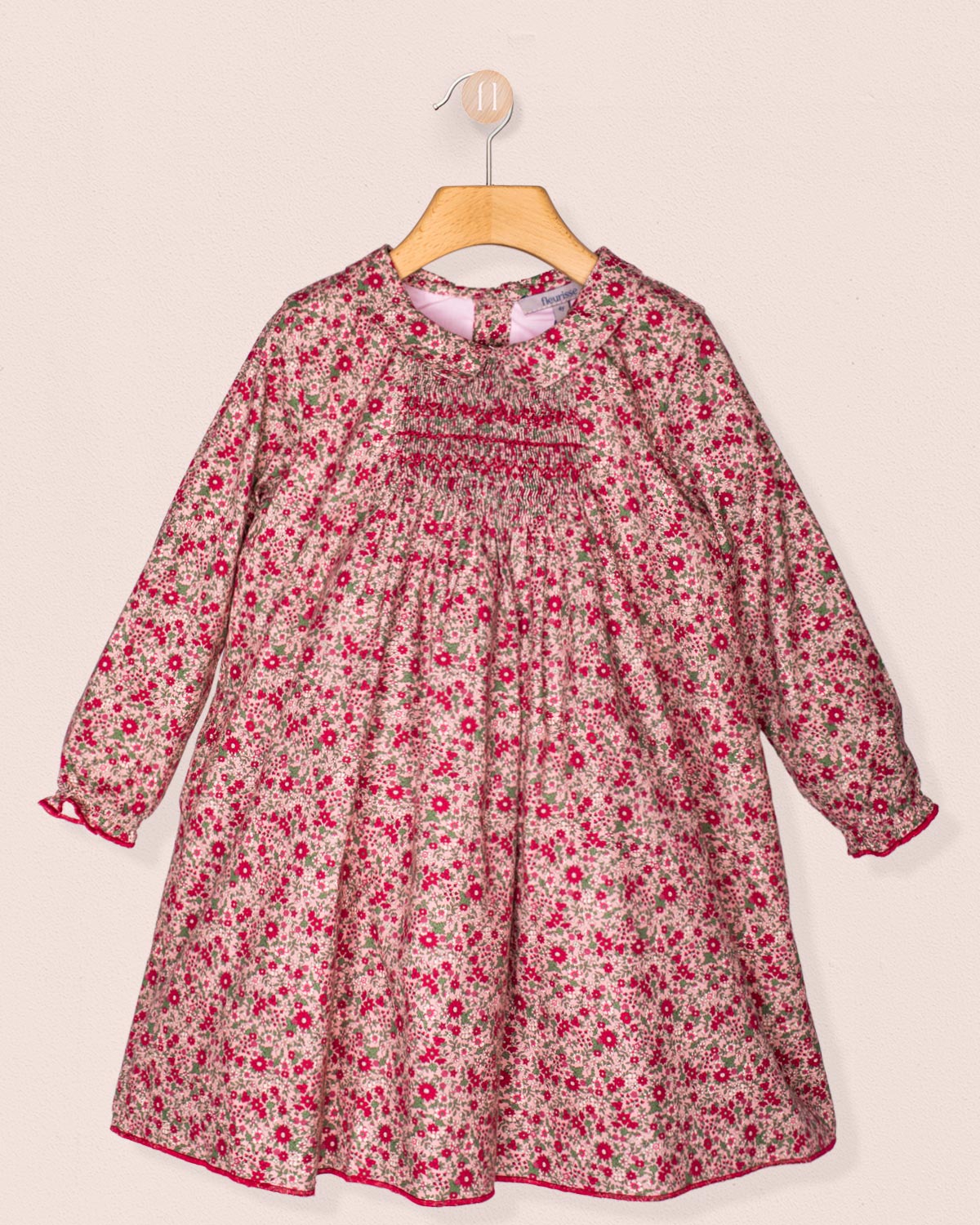 Front view of the Elizabeth Mini Mauve Floral Twill Dress hanging on a wooden hanger, showcasing its hand-smocked bodice, soft collar, and delicate mauve floral print.