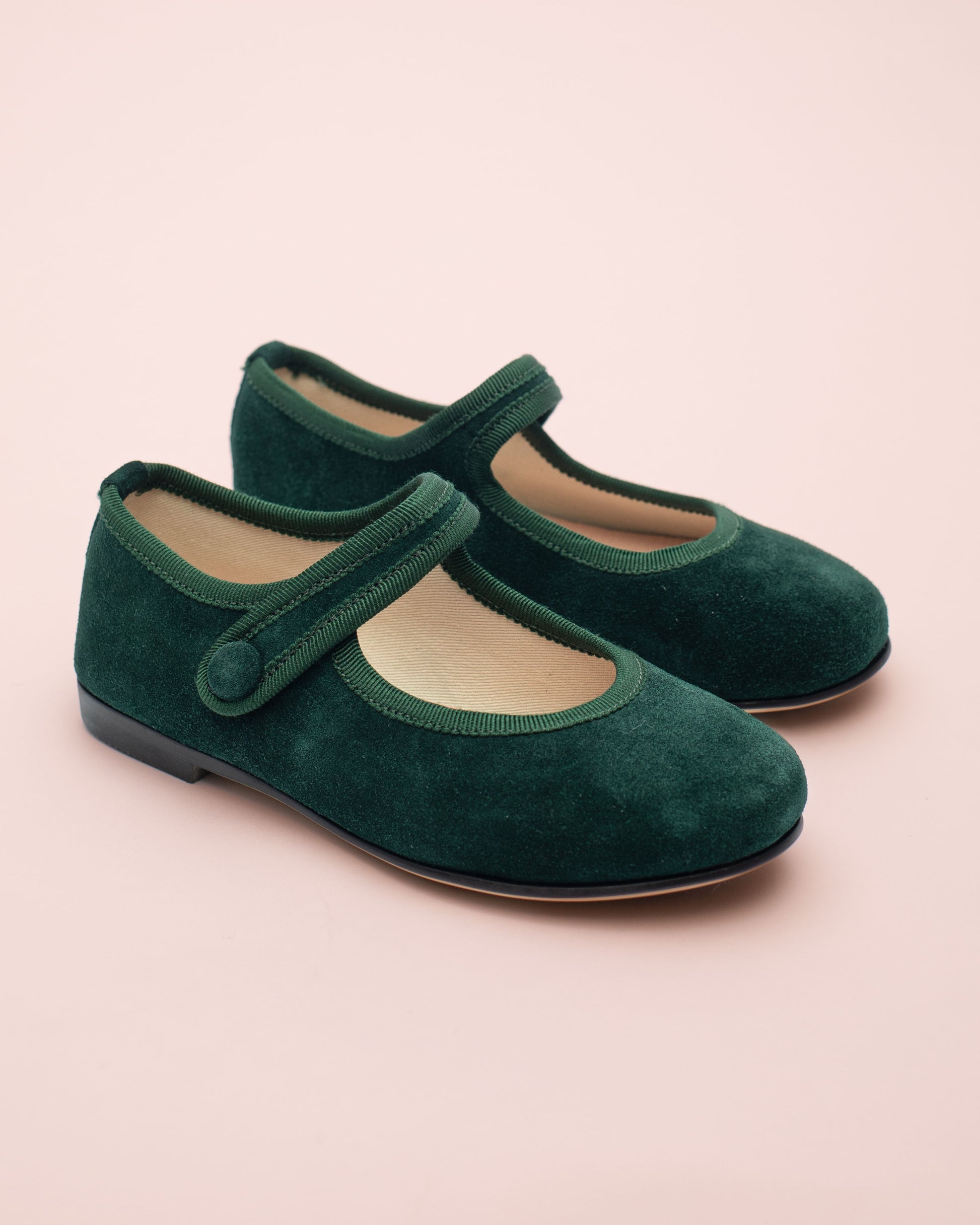 Pair of green suede shoes with a strap on a light pink background