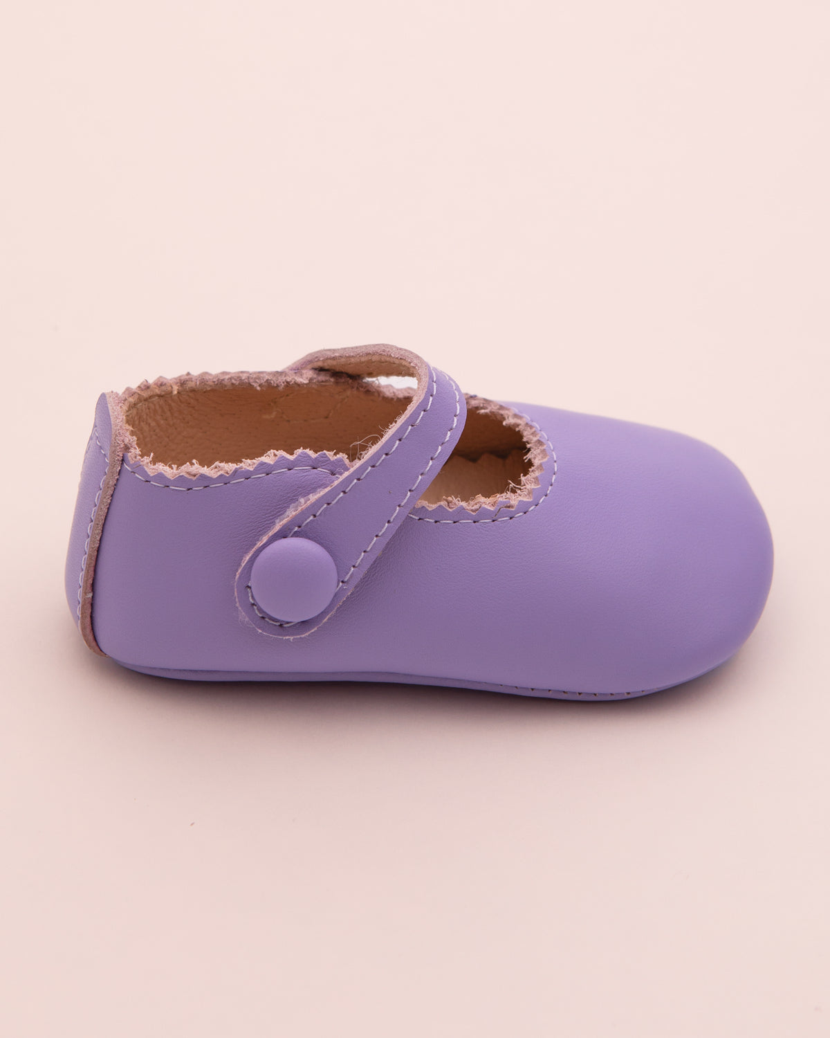 Side view of a lavender leather baby crib shoe with scalloped edge and velcro strap, shown against a pale pink backdrop.