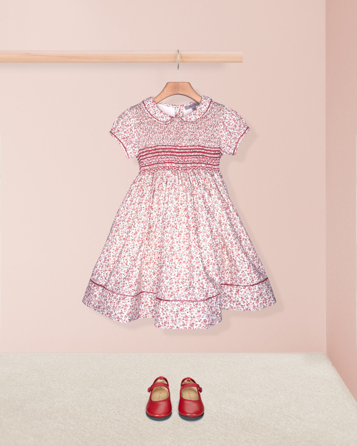 Pink floral dress with red trim on a hanger against a pink wall, accompanied by red shoes on the floor.