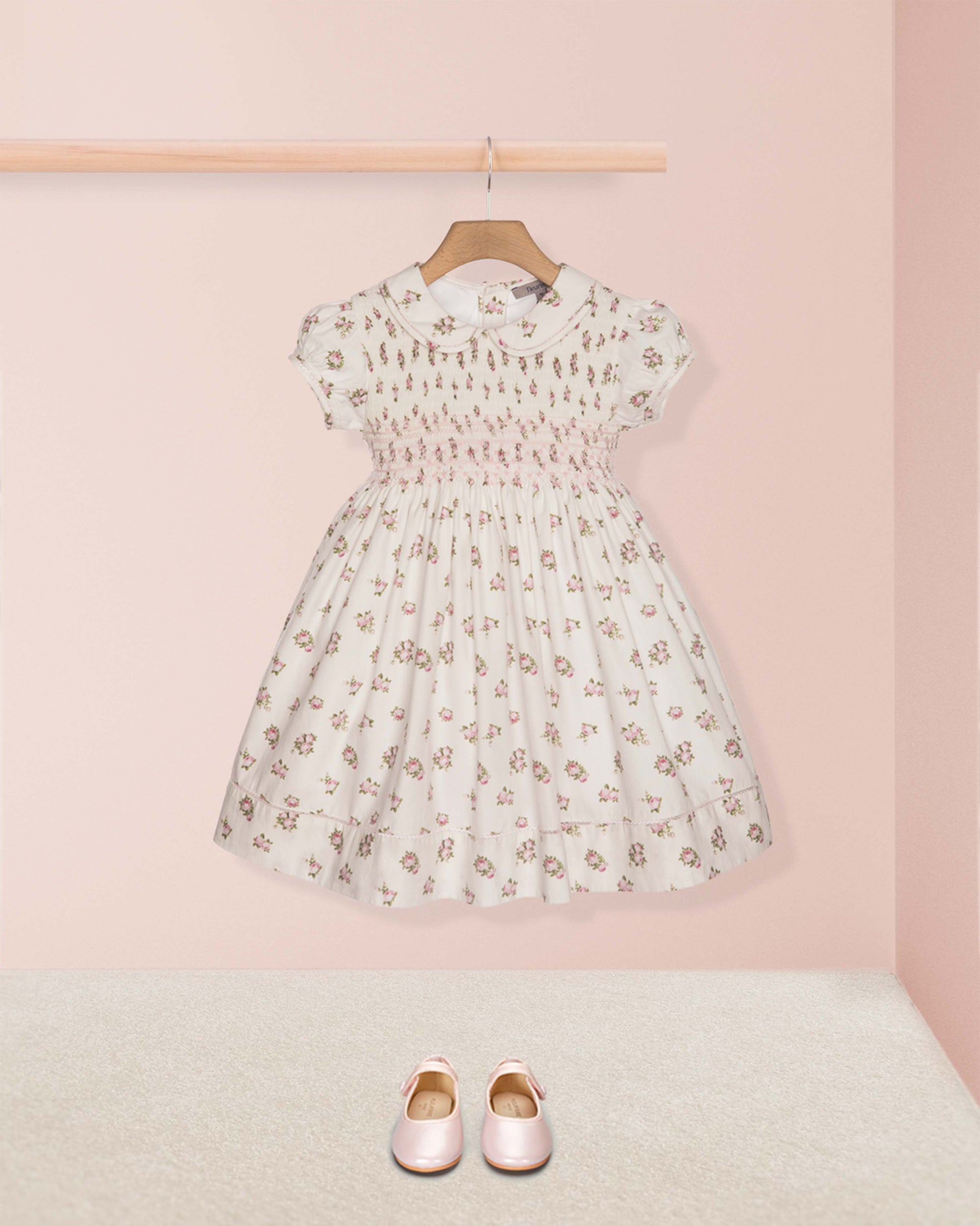 Floral dress on a hanger with pink shoes on the floor against a light pink wall.