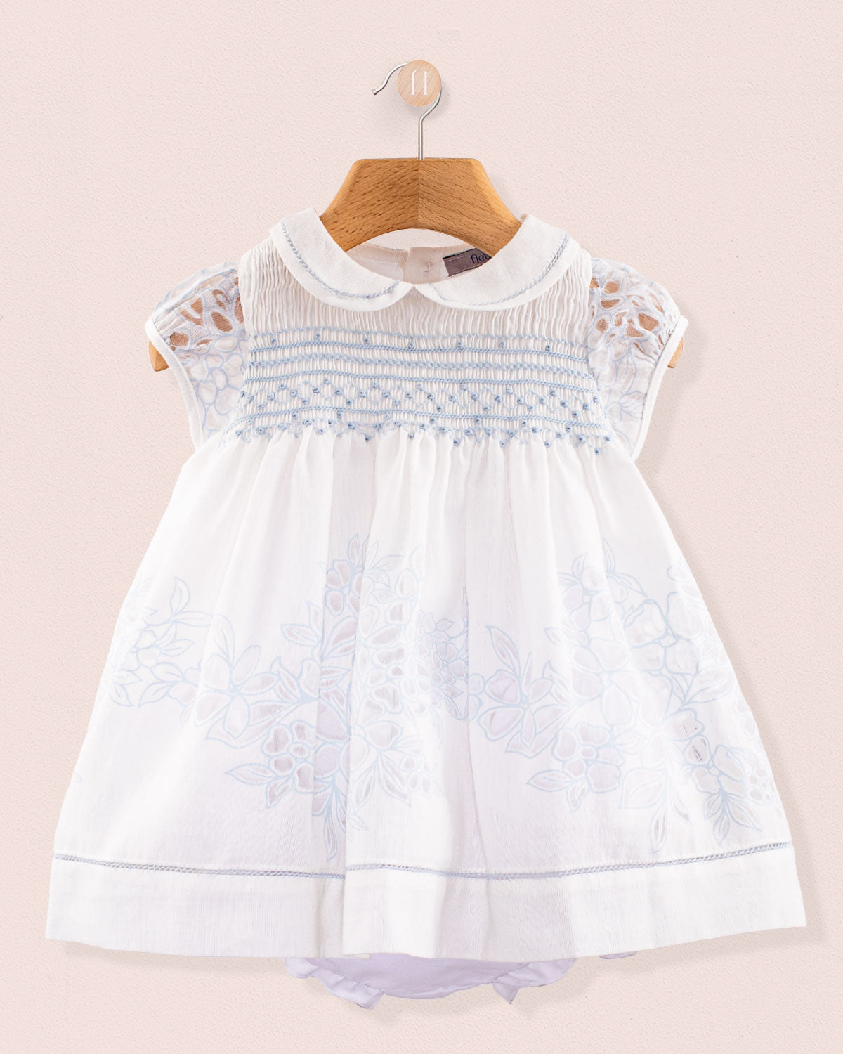 Front view of Baby Emmy Doppia Blue Smock Dress Set hanging on a wooden hanger showing blue hand smocking and floral embroidery on white cotton fabric