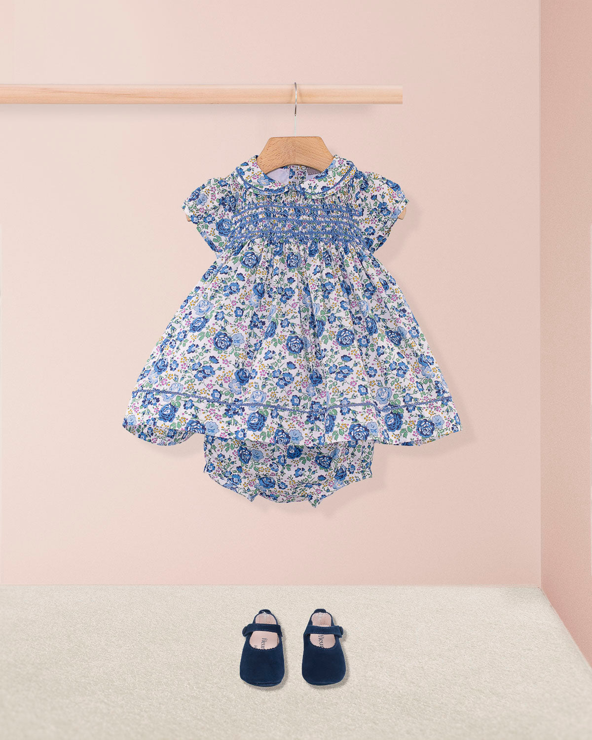 Baby Emmy Liberty Blue Rosebud Mini Dress Set with smocked bodice, entre-deux stitching, matching bloomers, and navy Mary Jane shoes on a pink backdrop.