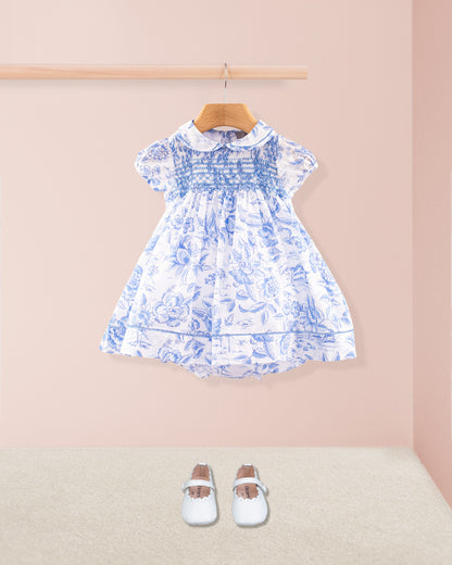 Full front view of the dress set displayed against a pink wall and cream carpet, paired with white baby Mary Jane shoes. The smocked blue bodice, floral skirt, and lace detailing are clearly visible.