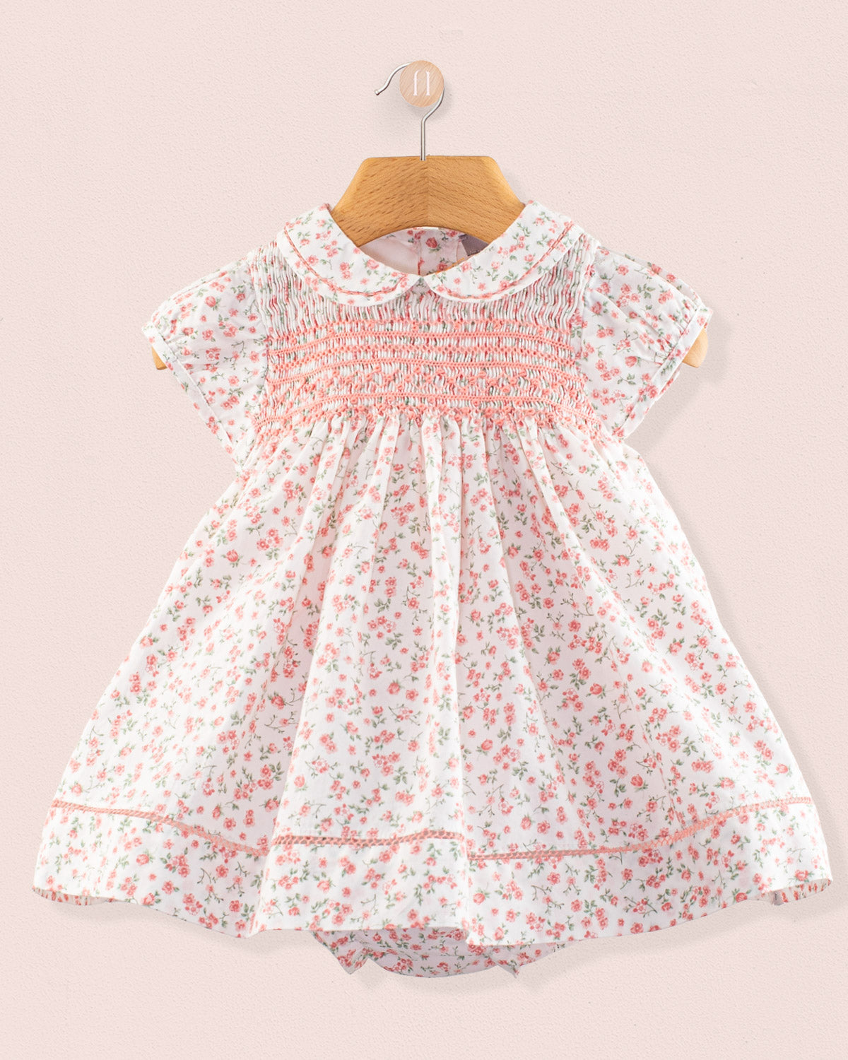 Front view of the Baby Emmy Elegant Pink Floral Dress Set hanging on a wooden hanger, featuring a smocked bodice, floral print, Peter Pan collar, and lace trim. Matching bloomers partially visible below.
