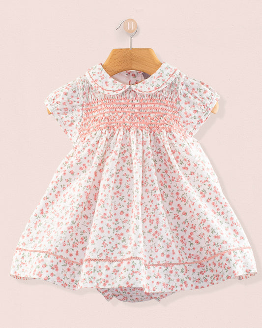 Front view of the Baby Emmy Elegant Pink Floral Dress Set hanging on a wooden hanger, featuring a smocked bodice, floral print, Peter Pan collar, and lace trim. Matching bloomers partially visible below.