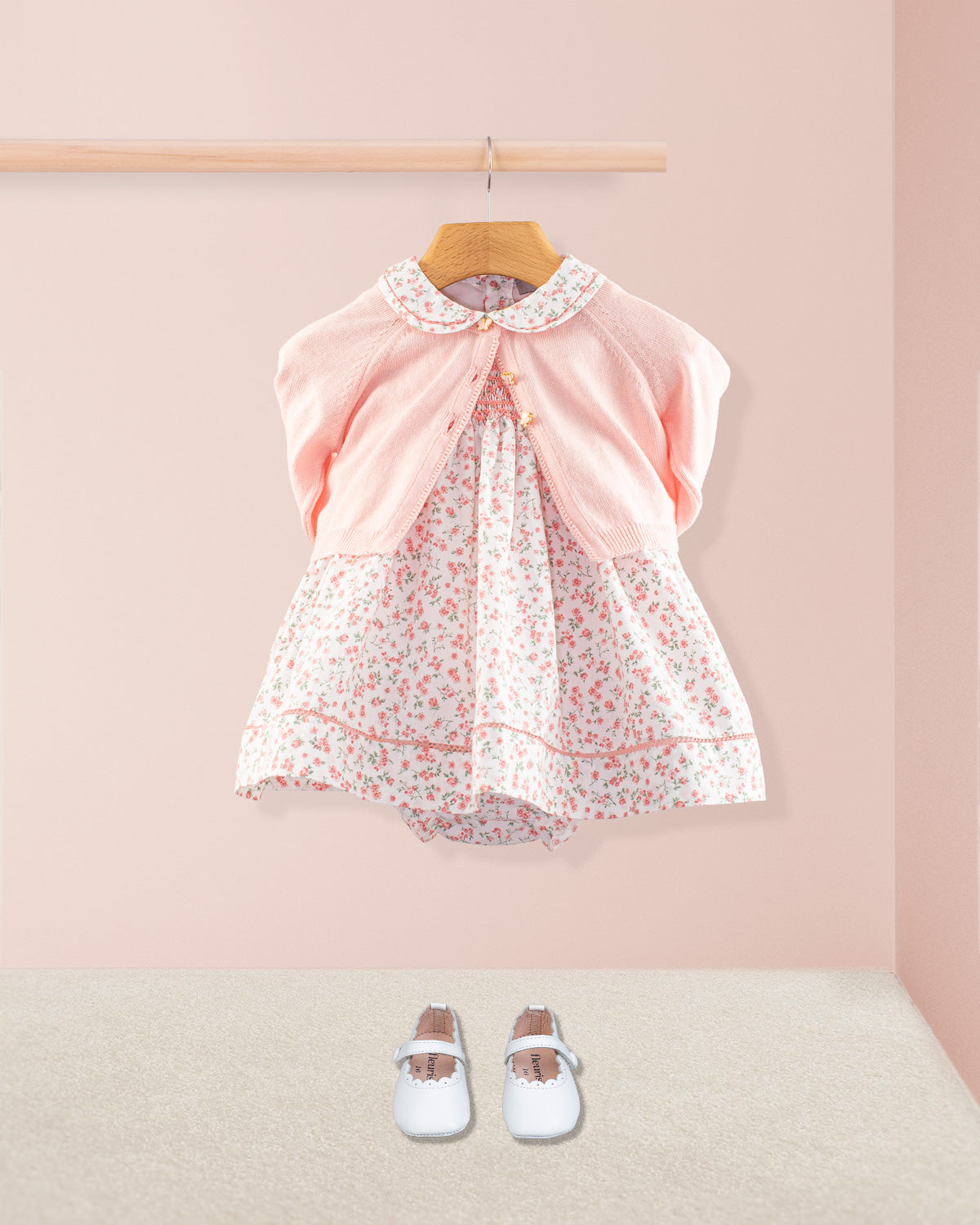 The Baby Emmy Pink Floral Dress styled with a petal pink knit cardigan and paired with white baby Mary Jane shoes. Dress is displayed hanging on a rod against a blush pink background.