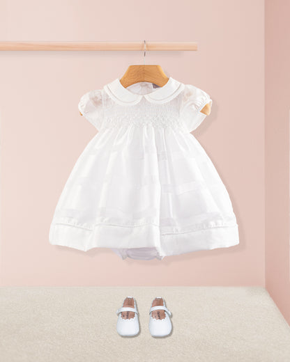 Baby Emmy White Chiffon Dress styled with soft white crib shoes placed on the floor below. The dress hangs on a wooden rod against a blush pink background with neutral carpet underneath.