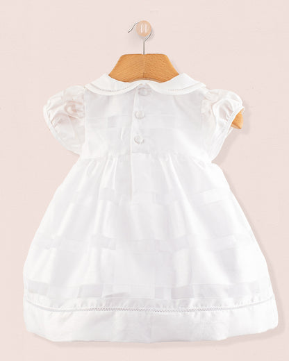 Back view of the Baby Emmy White Chiffon Dress on a wooden hanger. Features include three back buttons and a gently flared skirt, displayed against a light pink background