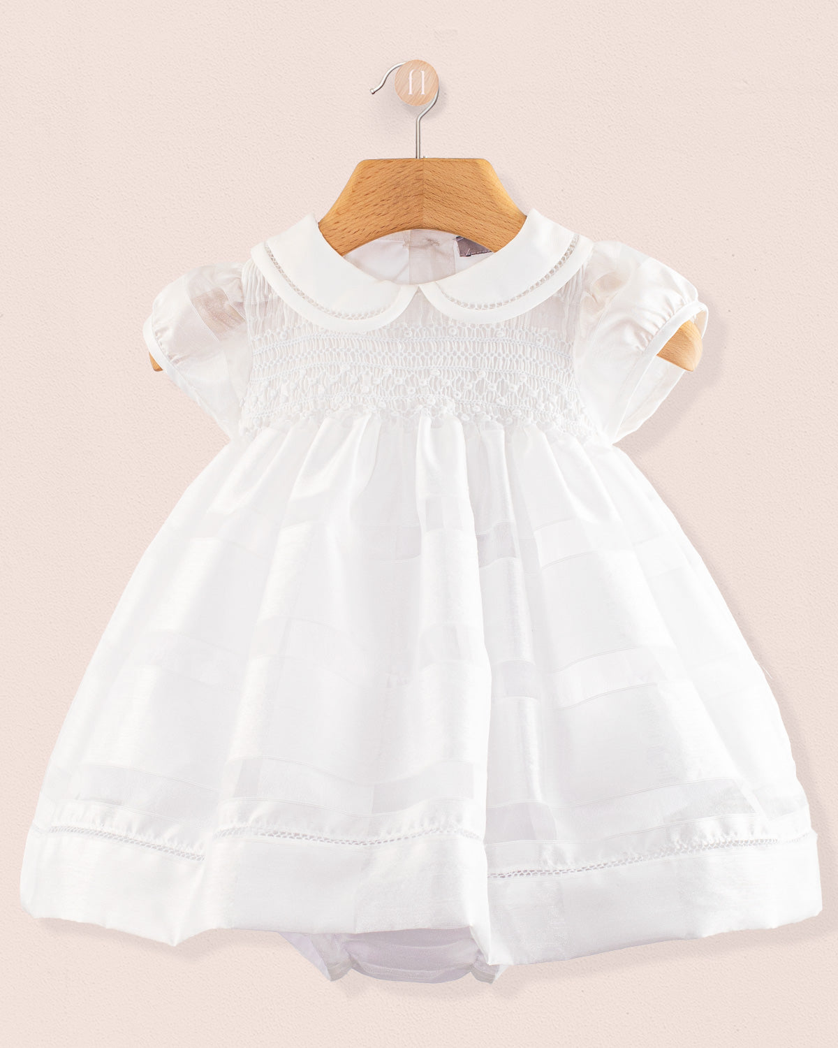 Front view of the Baby Emmy White Chiffon Dress hanging on a wooden hanger. The dress features short puff sleeves, a Peter Pan collar, and a fully smocked bodice with lace trim, displayed against a soft pink background.