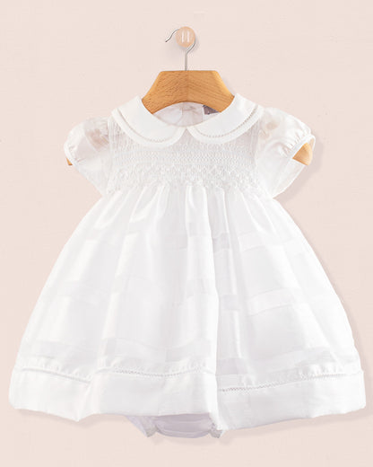 Front view of the Baby Emmy White Chiffon Dress hanging on a wooden hanger. The dress features short puff sleeves, a Peter Pan collar, and a fully smocked bodice with lace trim, displayed against a soft pink background.