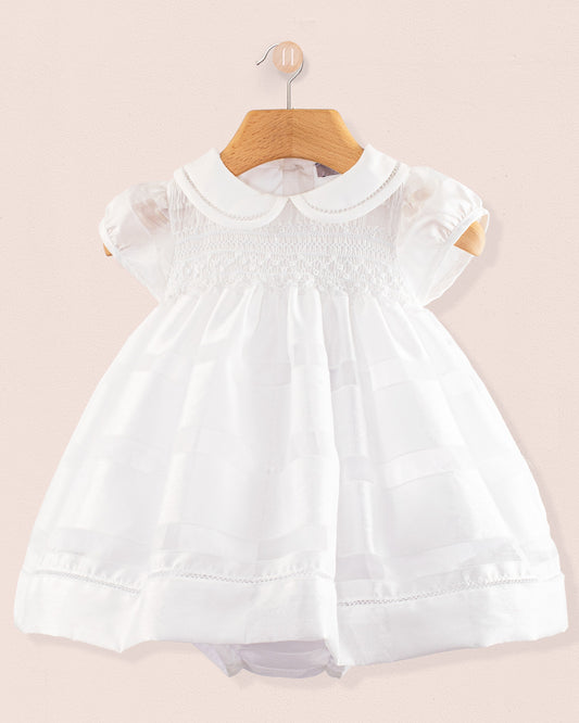 Front view of the Baby Emmy White Chiffon Dress hanging on a wooden hanger. The dress features short puff sleeves, a Peter Pan collar, and a fully smocked bodice with lace trim, displayed against a soft pink background.
