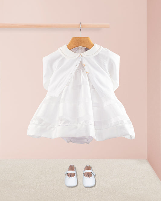 Baby Emmy White Chiffon Dress styled with a matching white cardigan and soft white crib shoes. The dress set hangs from a light wood dowel against a blush pink background with beige carpeted floor.