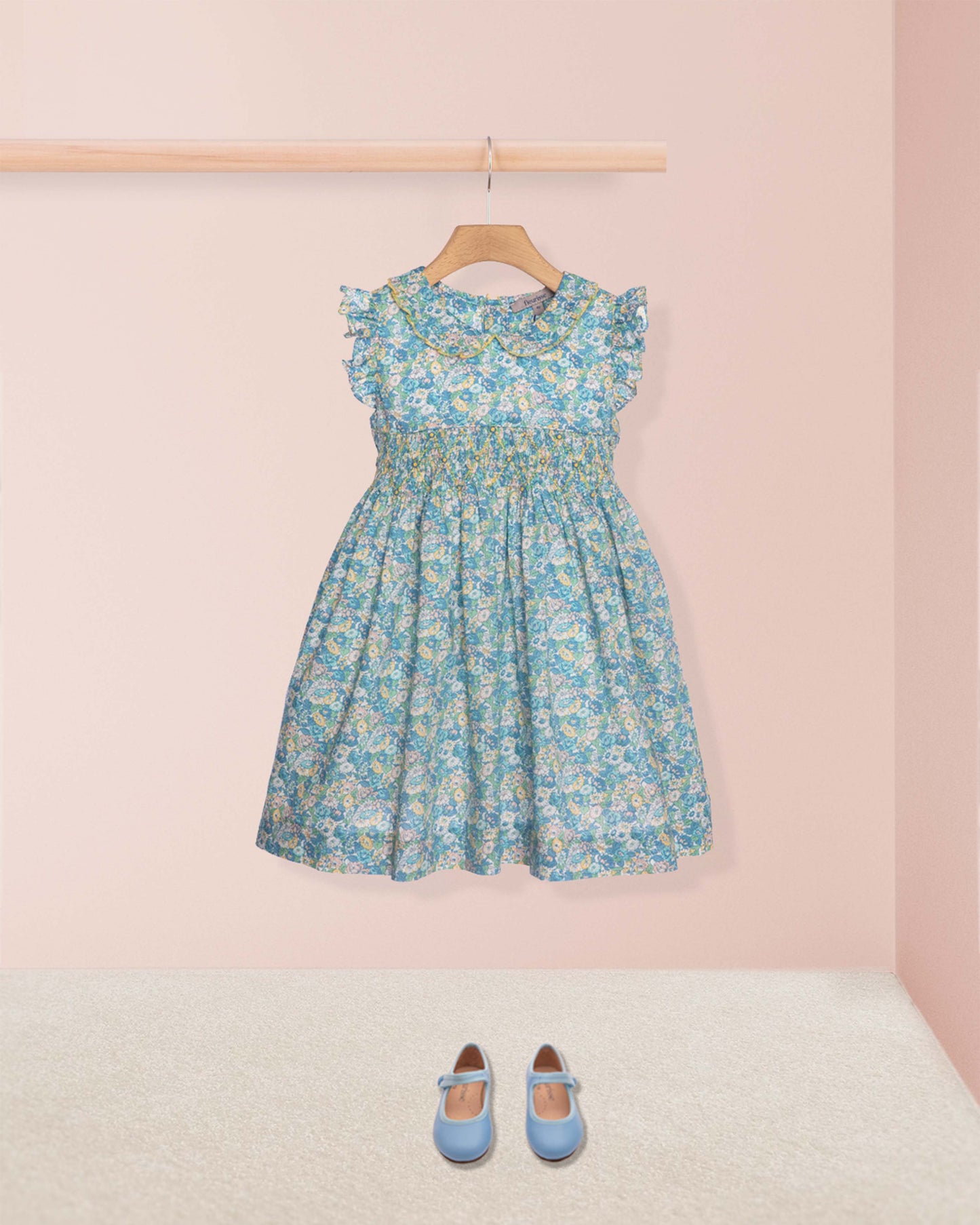 Floral dress hanging on a rack with blue shoes on the floor against a pink wall.