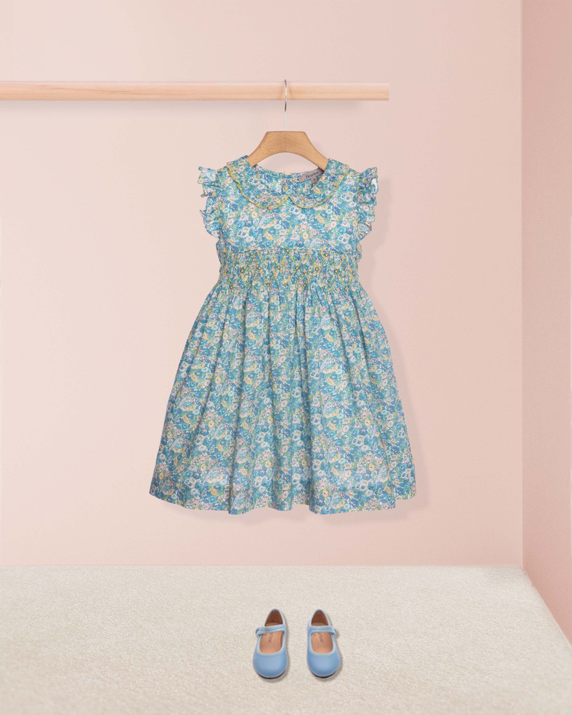 Floral dress hanging on a rack with blue shoes on the floor against a pink wall.