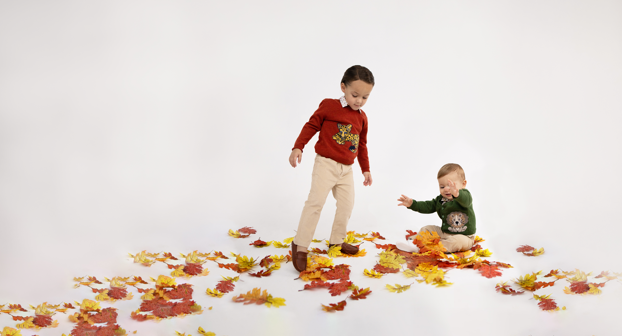 2 boys wearing sweaters with a leopard and a dog, kicking leaves in a studiolike white backdrop.