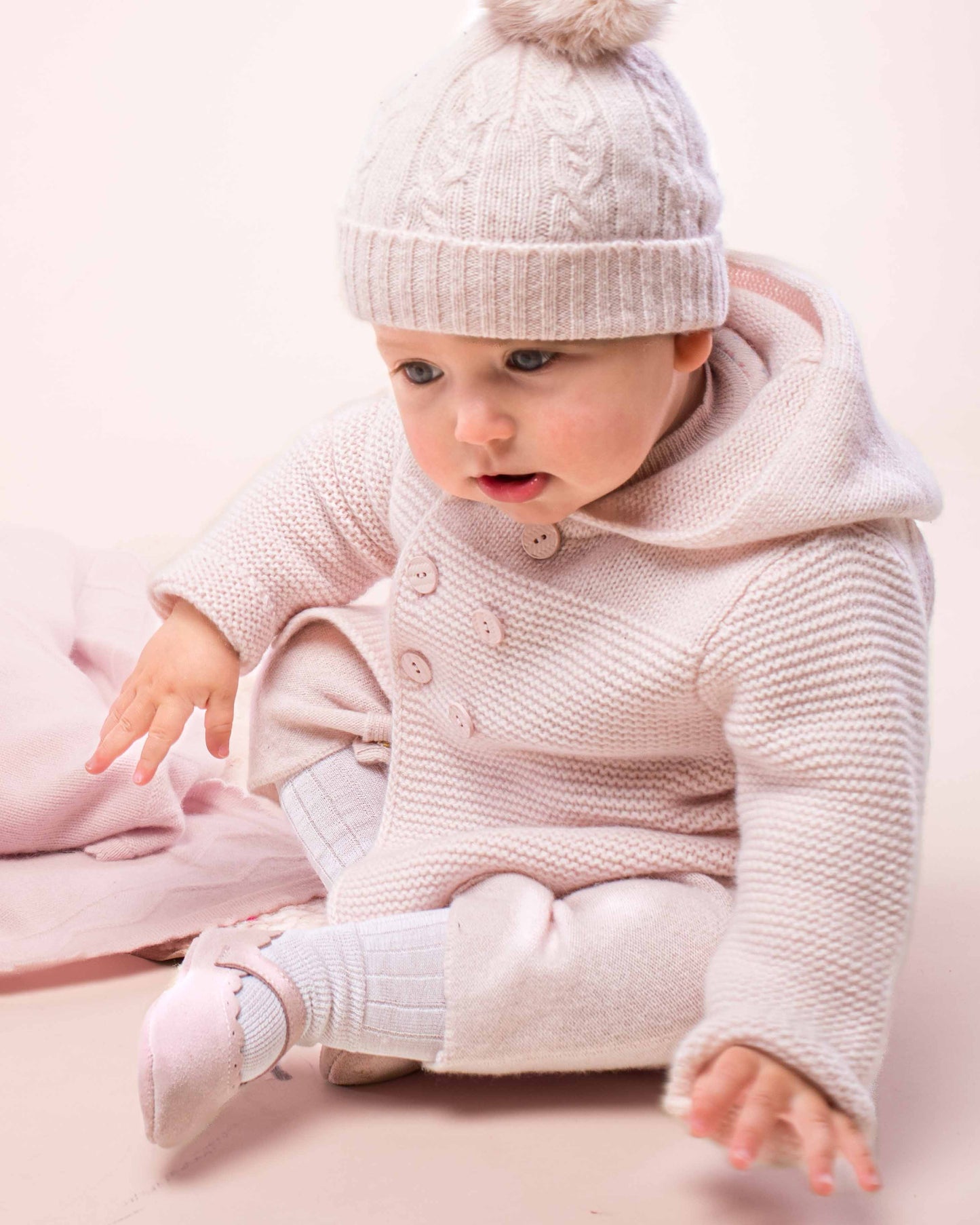 Baby sitting on the floor, looking down, wearing the Chamonix Powder Pink Cashmere Hooded Jacket and a matching hat and pants, showing the front of the jacket