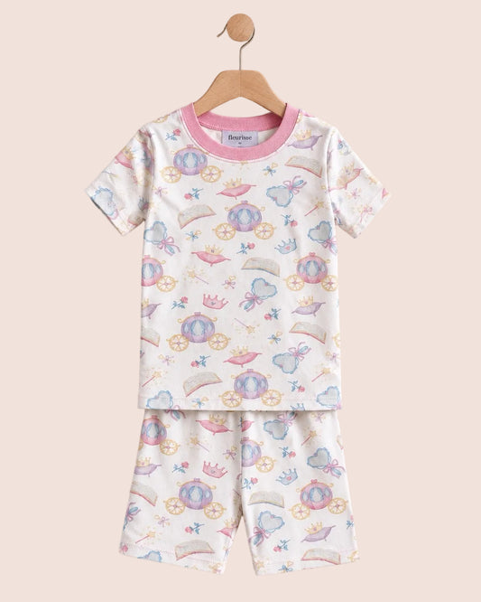 A two-piece baby pajama set featuring a short-sleeve top and matching shorts, hanging on a wooden hanger against a solid pale pink background. The pajamas have a white base with a colorful print of whimsical fairies, castles, and rainbows, and are finished with a pink ribbed collar.