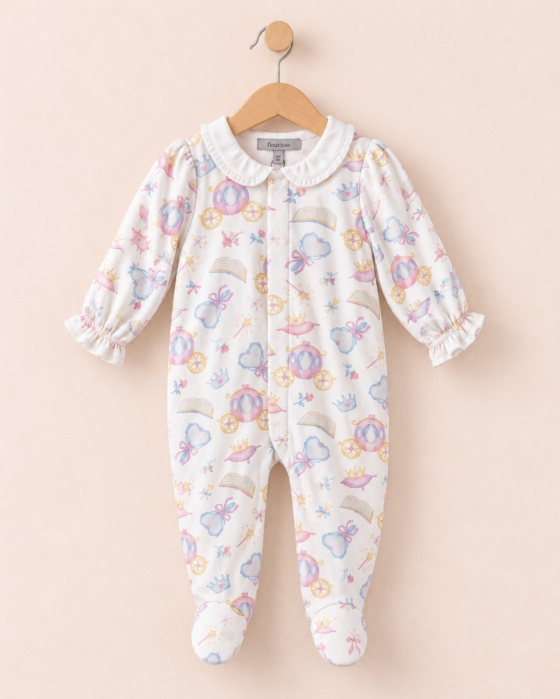 a one-piece baby onesie hanging on a wooden hanger against a solid pale pink background. The onesie has a white base with a colorful print of whimsical fairies, castles, and rainbows, and is finished with a white collar.