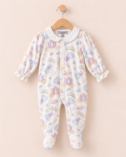a one-piece baby onesie hanging on a wooden hanger against a solid pale pink background. The onesie has a white base with a colorful print of whimsical fairies, castles, and rainbows, and is finished with a white collar.